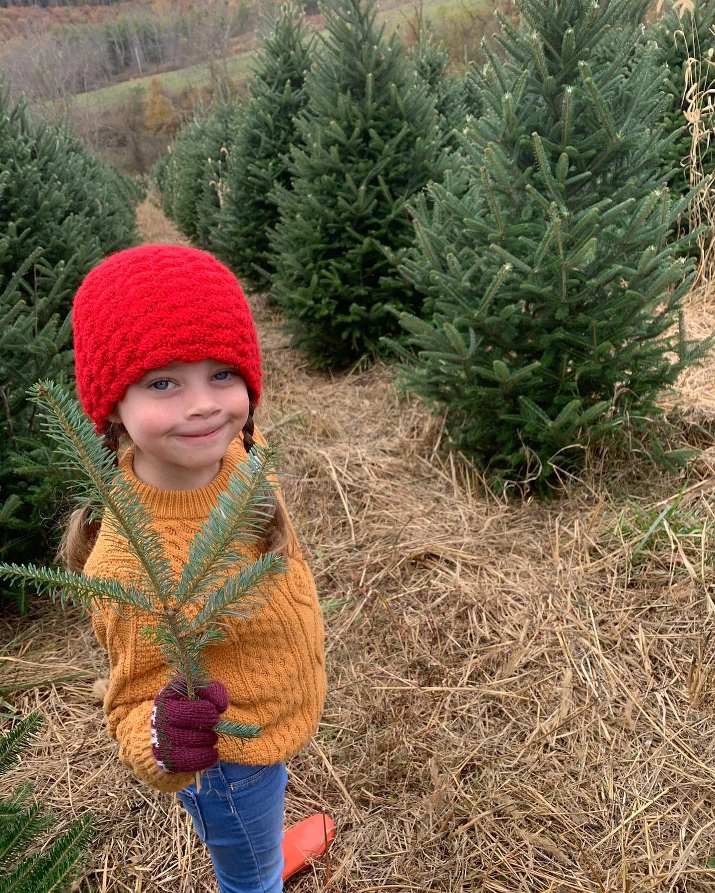 A girl in a red knit hat and mustard sweater holding a Fraser Fir branch in the hay