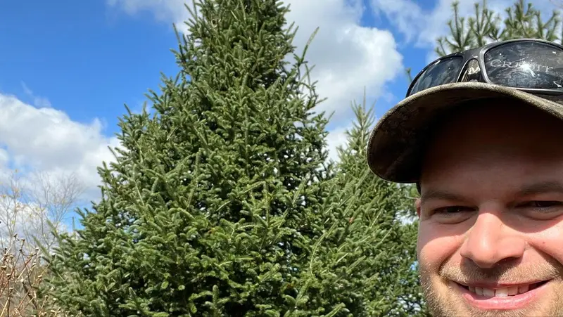 A grinning selfie in a farm cap beside the tall Fraser Fir chosen for the Executive Mansion