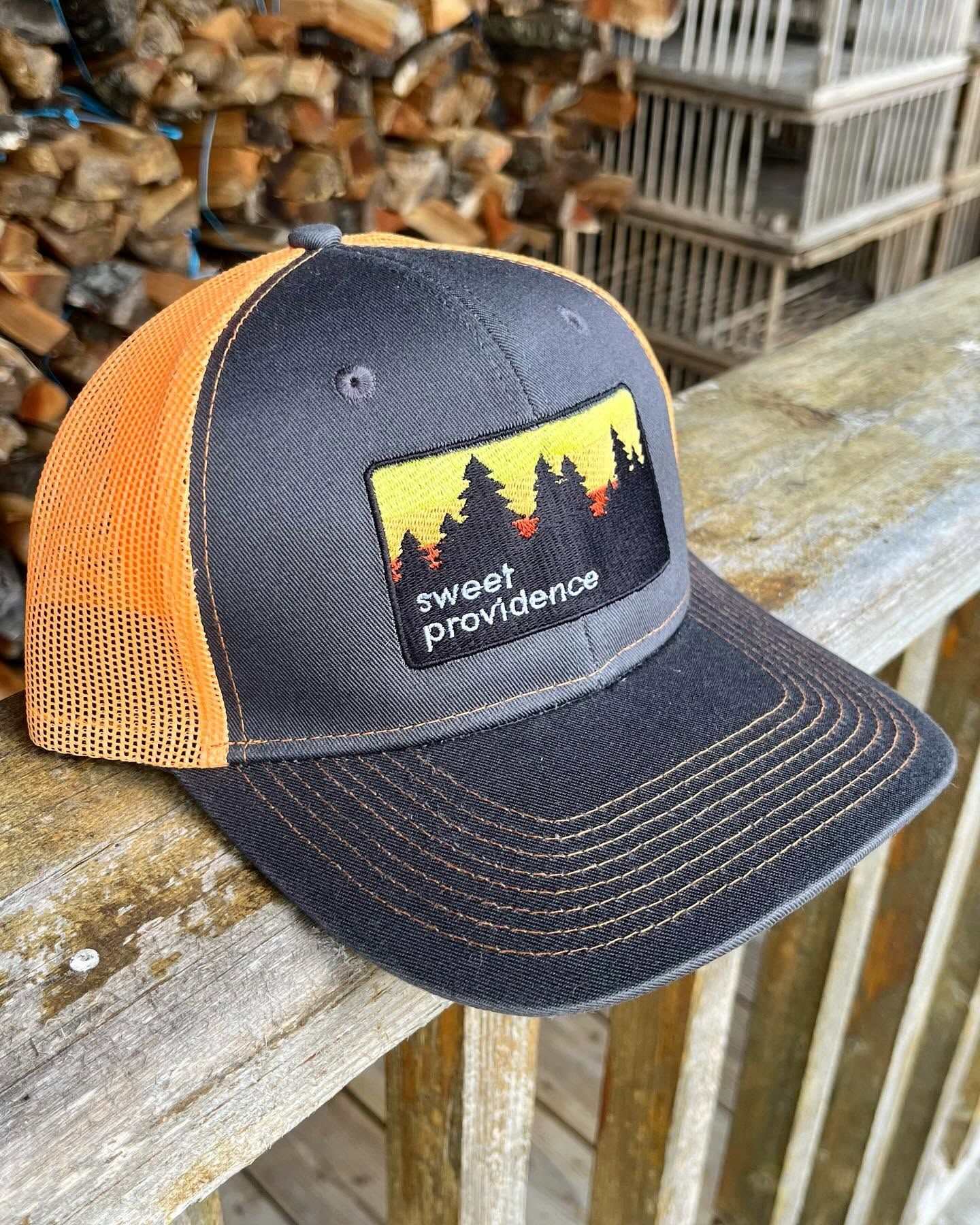 A charcoal and orange trucker cap with a sunset-over-the-trees patch that reads Sweet Providence